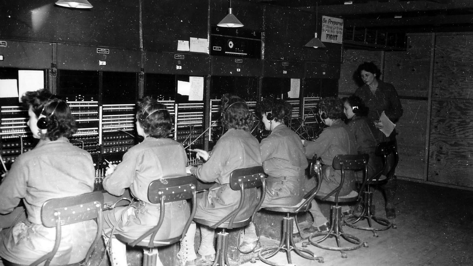 A row of telephone operators working a switchboard while a supervisor checks the log